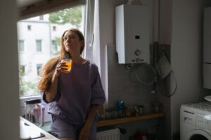 A woman holding a drink and looking thoughtfully through the camera. A boiler is on the wall next to her.